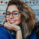 A young woman wearing glasses and denim, smiling in an indoor setting.